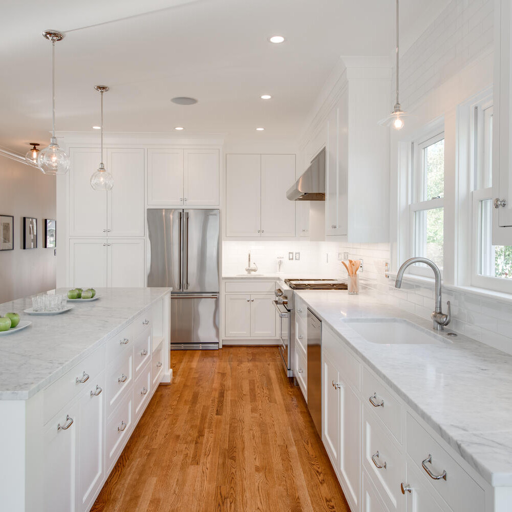 A bright, modern kitchen with white cabinets, marble countertops, and wooden floors. Three pendant lights hang above the island, which has green apples on it. Stainless steel appliances are visible, and a window allows natural light to enter.