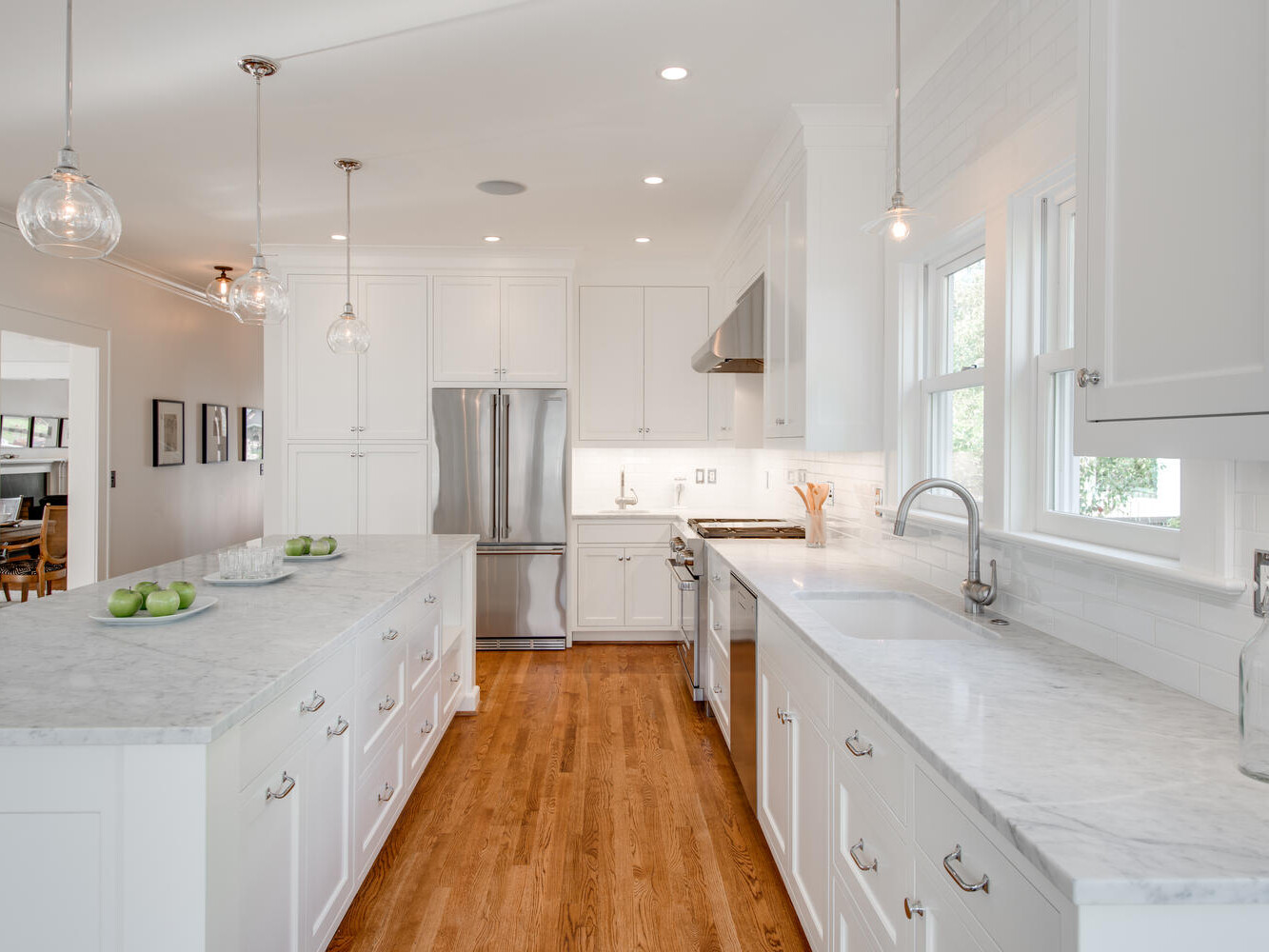 A bright, modern kitchen with white cabinets, marble countertops, and wooden floors. Three pendant lights hang above the island, which has green apples on it. Stainless steel appliances are visible, and a window allows natural light to enter.