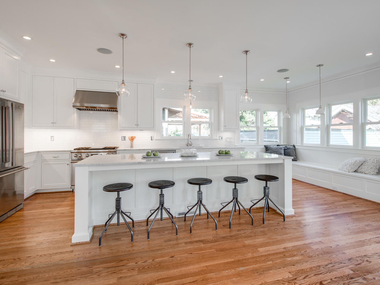 A bright, modern kitchen with hardwood floors, white cabinetry, and a central island. The island has black stools and pendant lights above. Stainless steel appliances are visible, and a window seat with cushions is on the right.