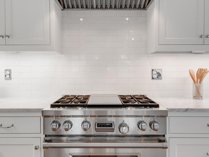 A modern kitchen featuring a stainless steel stove with a large oven and six burners. Above is a sleek range hood. The white subway tile backsplash and light-colored cabinets create a bright and clean look. A set of wooden utensils is on the right.