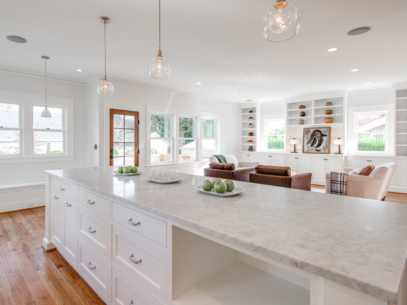 Spacious open-concept kitchen and living area with a large marble island. The kitchen features white cabinetry, pendant lights, and a wooden floor. The living space has brown leather sofas and built-in shelves. Large windows allow natural light.