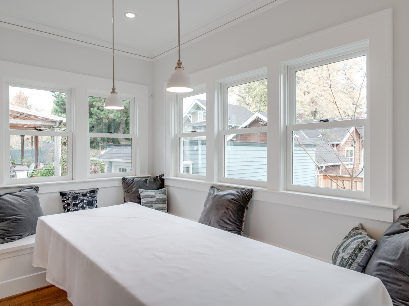 A bright dining nook with a long white table, surrounded by gray and patterned cushions on built-in benches. Three large windows offer views of neighboring houses and trees. Two pendant lights hang from the ceiling, illuminating the space.
