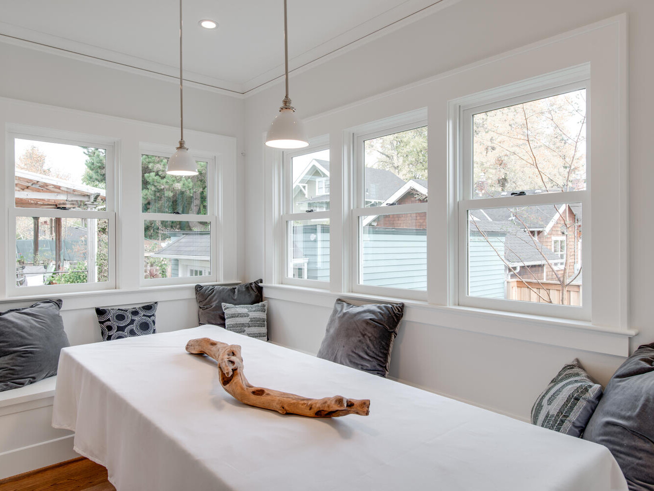 A cozy dining nook with a long table covered by a white tablecloth and decorated with a wooden centerpiece. The seating area has dark gray cushions and is surrounded by large windows with a view of neighboring houses and trees.