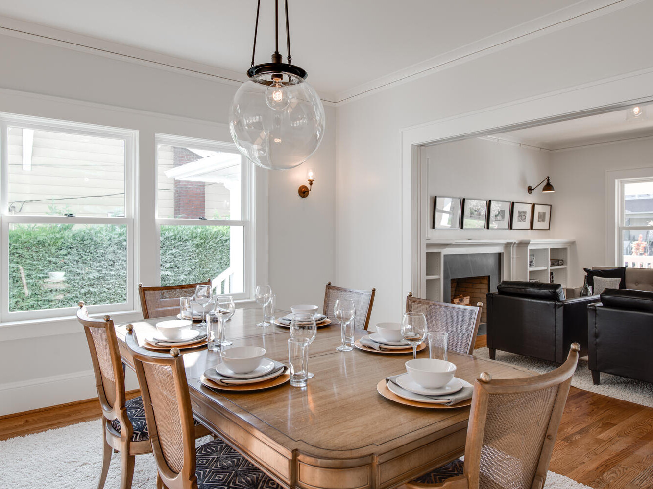 A bright dining room with a wooden table set for six, featuring plates, bowls, and glasses. Above the table hangs a spherical pendant light. The room has large windows, and an adjacent living area with black chairs and a fireplace is visible.