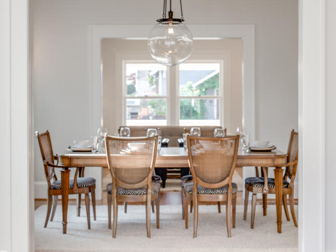 A dining room with a wooden table set with plates, glasses, and chairs with woven backs. A large glass light fixture hangs above the table. Visible through a doorway, the room has large windows with a view of trees. A decorative bowl is in the foreground.