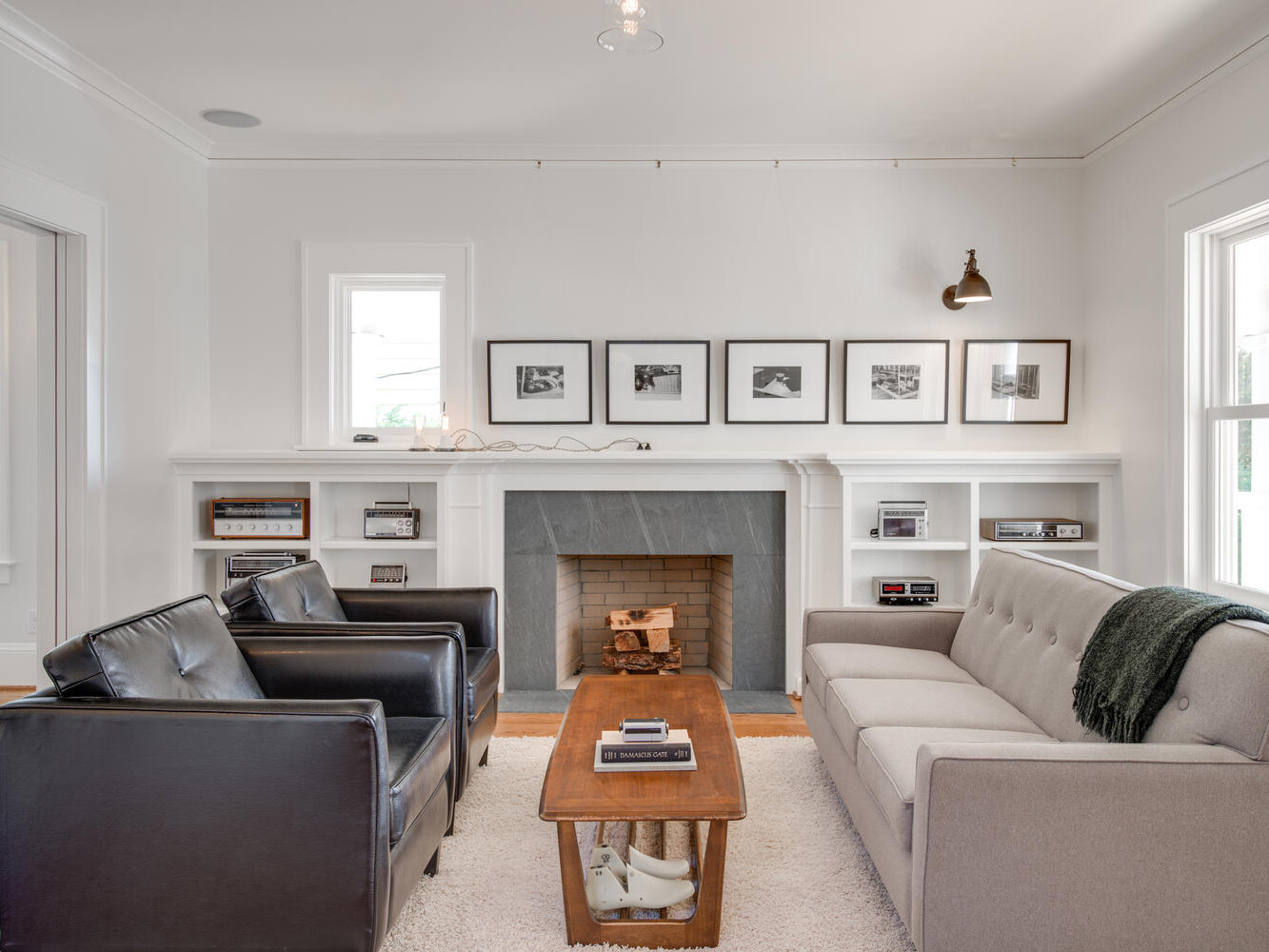 A cozy living room features a gray sofa, two black armchairs, and a wooden coffee table. A fireplace is adorned with framed pictures above it. Light streams in through large windows, highlighting the modern, minimalist decor.