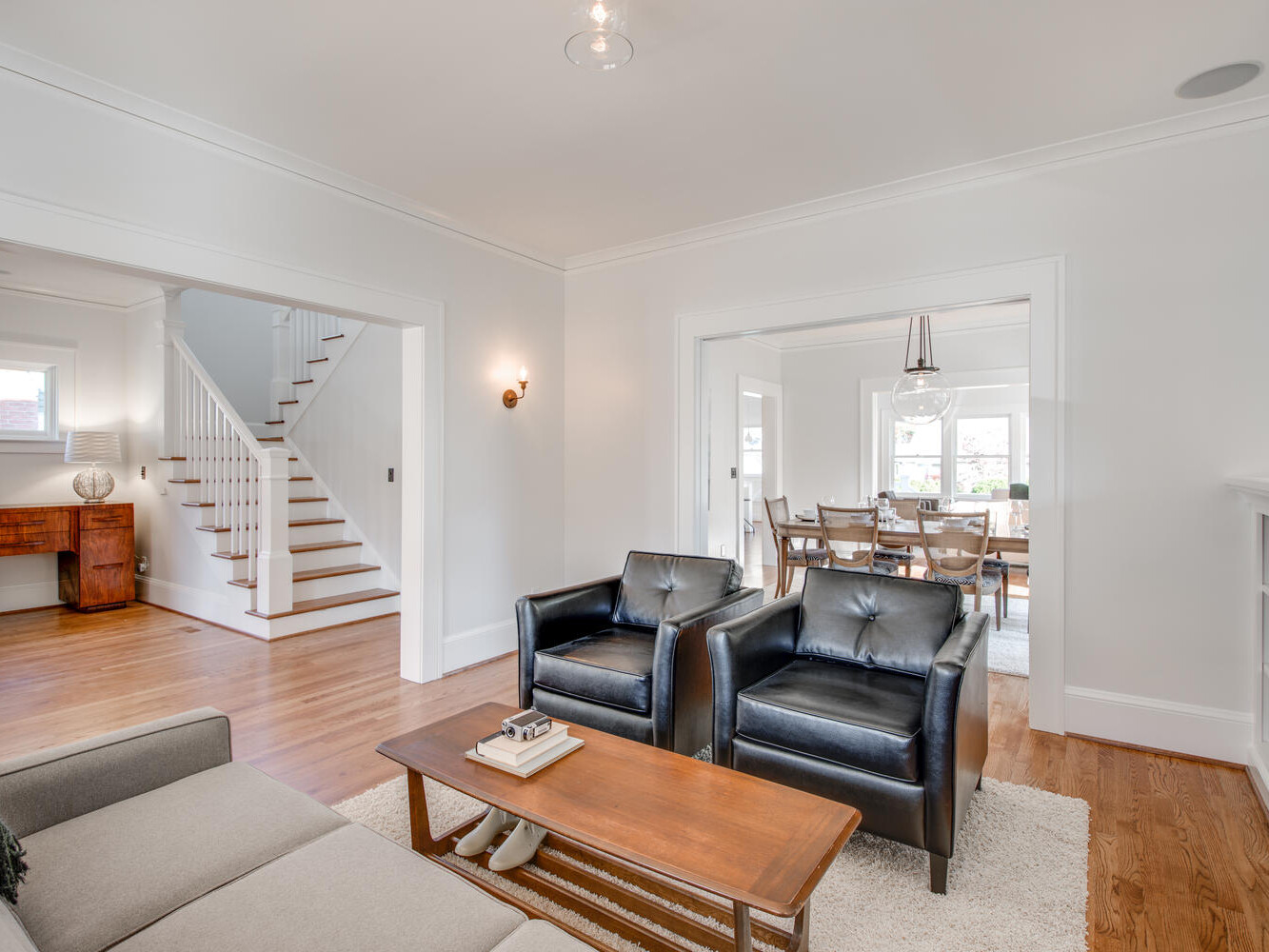 A bright living room with two black leather chairs, a gray sofa, and a wooden coffee table. The room opens to a dining area with a table and chairs. Wooden floors and a white staircase are visible in the background.