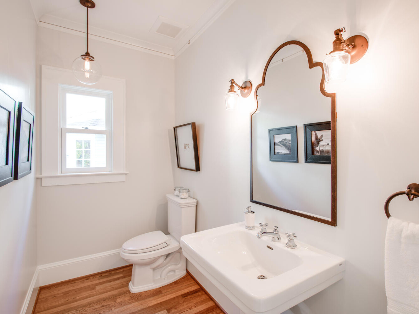 A bright, narrow bathroom with a large mirror, two wall-mounted lights, a white sink, and a toilet. Wooden flooring complements the neutral walls, and framed pictures are hung on the walls. A small window lets in natural light.