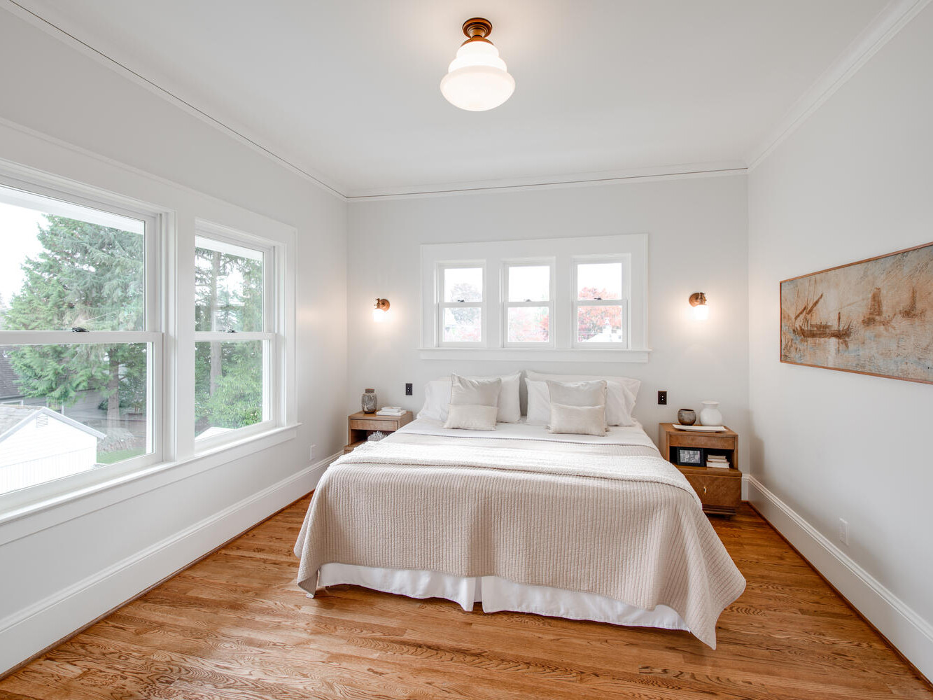 A bright, minimalist bedroom features a bed with light-colored bedding and pillows. Large windows on two sides let in natural light, highlighting the wooden floor. A wall painting and bedside tables complete the cozy setting.