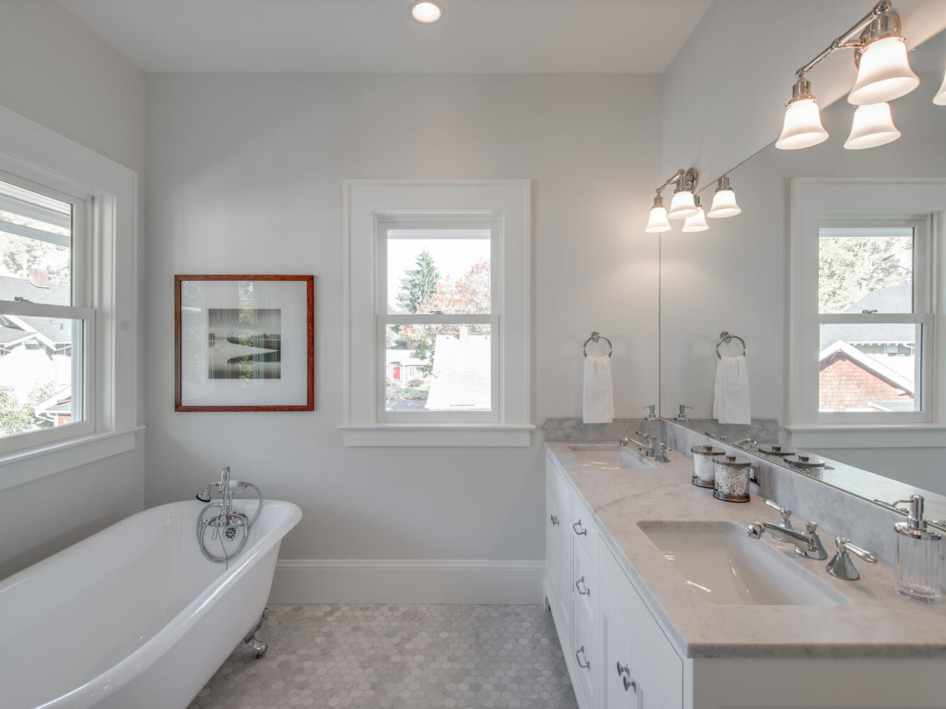A bright bathroom with a white clawfoot bathtub beneath a window, a double-sink vanity with a marble countertop, two mirrors, and wall-mounted lights. Gray hexagonal floor tiles and a framed artwork next to the window complete the space.