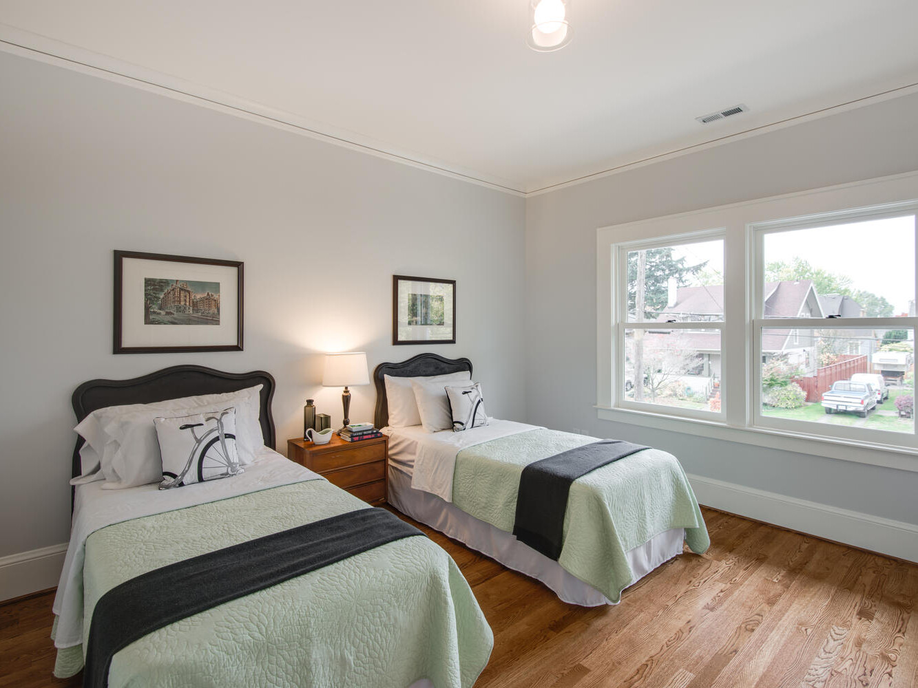 A tidy bedroom with two single beds featuring pale green and dark gray bedding. A wooden nightstand with a lamp and books is between the beds. Two framed pictures hang above the beds, and a window offers a view of a garden and houses outside.