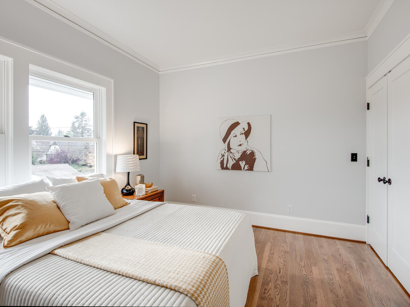 A bright bedroom with light gray walls and wooden flooring. The bed has white and mustard-yellow bedding. Theres a nightstand with a lamp and decor items. A large window lets in natural light, and a minimalist artwork hangs on the wall.