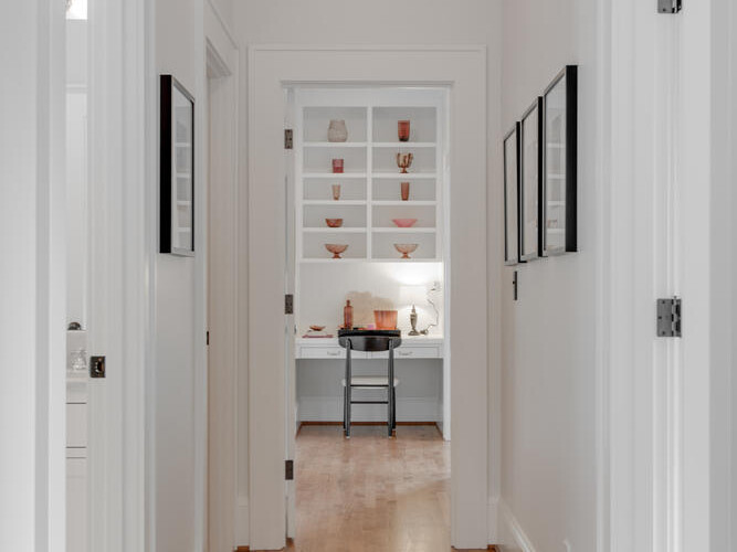 A narrow, white hallway with wooden floors leads to a bright room. Shelves with decorative items and a small table are visible at the end. Two doors are on each side of the hallway. A ceiling light illuminates the space.