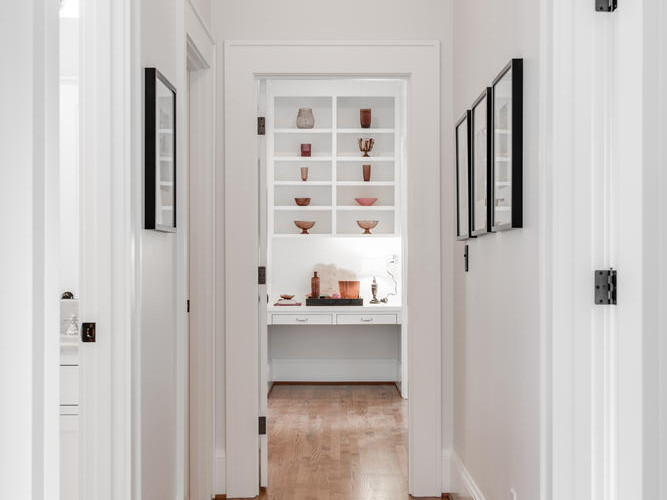 A bright, narrow hallway with wooden floors and white walls leads to a room with shelves displaying pottery. Framed artwork decorates the walls. A ceiling light fixture illuminates the space.