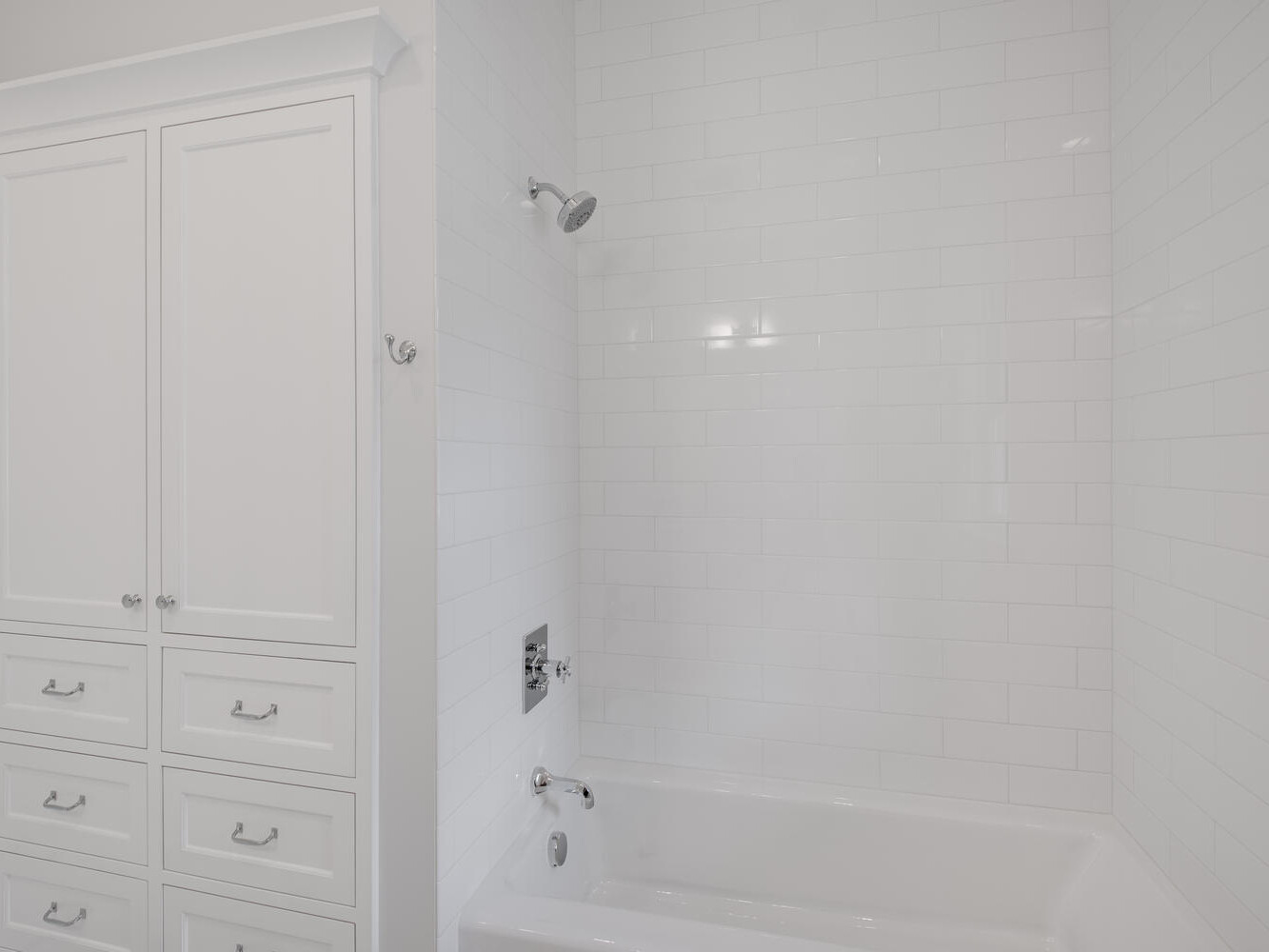 A clean, white bathroom features a bathtub with a wall-mounted showerhead. The walls are tiled, and there is a tall white cabinet with drawers on the left. The overall look is modern and minimalistic.