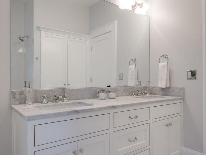 A modern bathroom with a large mirror above a white dual-sink vanity with marble countertop. Bright lights are mounted above the mirror, and white towels hang on a wall hook. The floor features grey and white hexagonal tiles.
