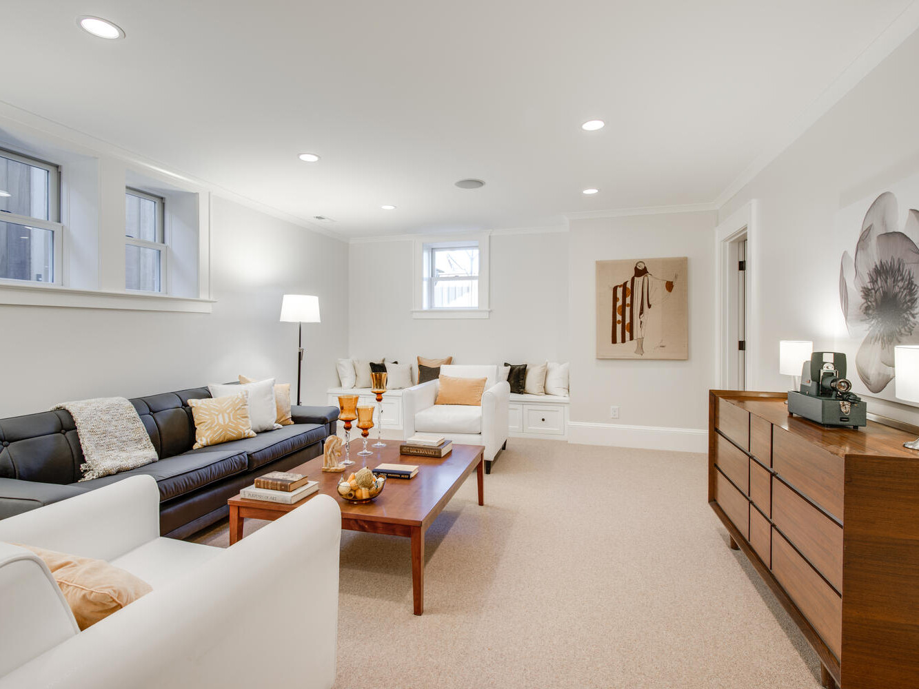 A cozy living room featuring a black and white sofa, a wooden coffee table, and a chest of drawers. The room is adorned with flower-themed artwork and various decorative pillows. Soft lighting creates a warm ambiance.