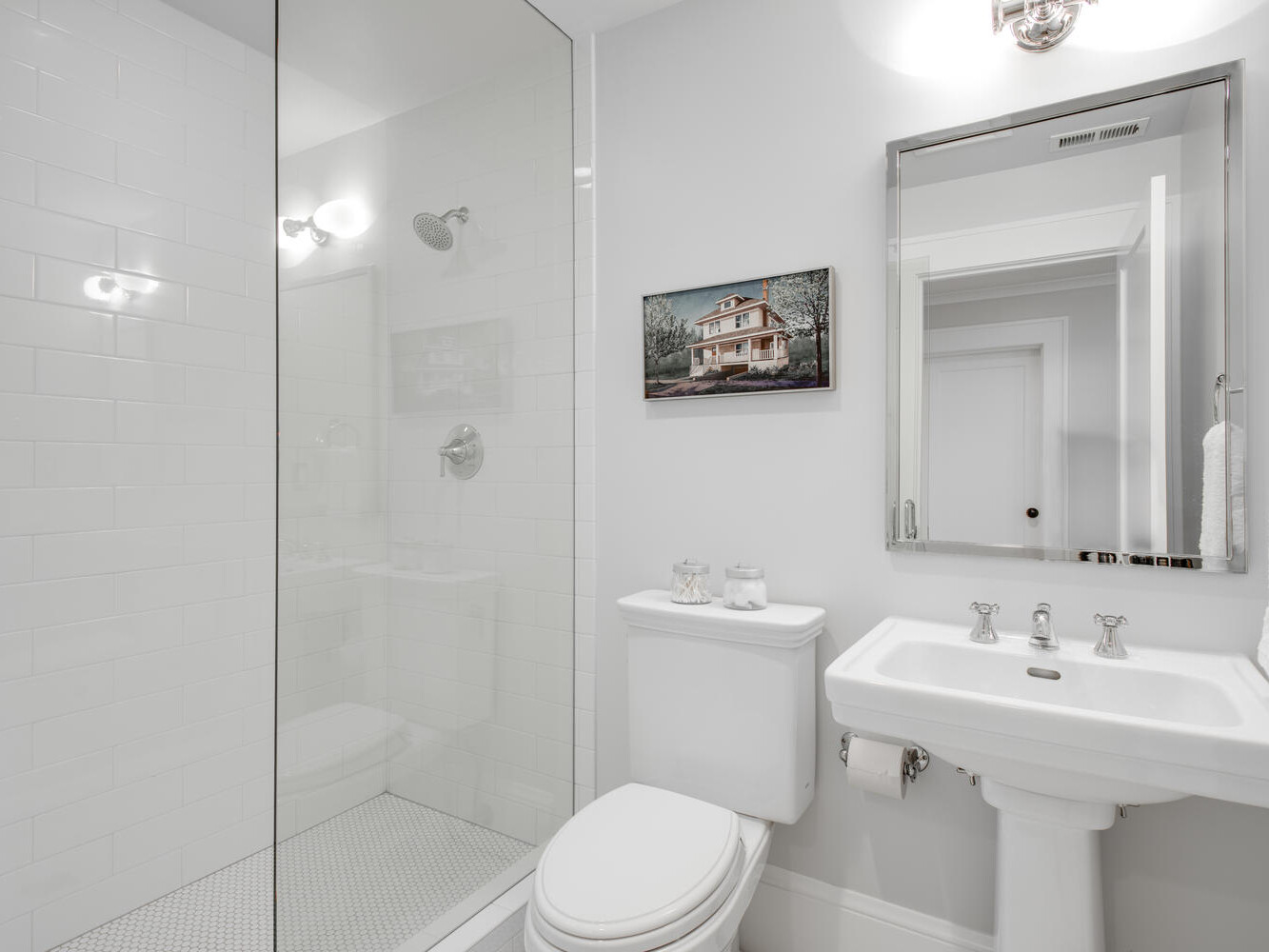 A modern bathroom with a glass-enclosed shower, white subway tiles, a pedestal sink, and a toilet. A rectangular mirror is above the sink, and a framed picture hangs on the wall. Neutral color palette with bright lighting.