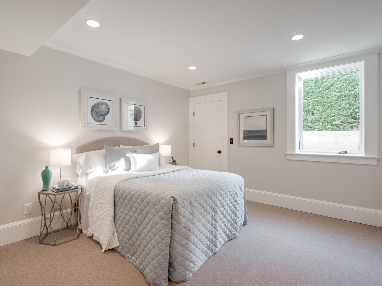 A serene bedroom with a double bed dressed in a light gray quilt and white pillows. Two framed seashell prints hang above the bed. Two bedside tables hold lamps. A window on the right side provides natural light. The walls and ceiling are white.