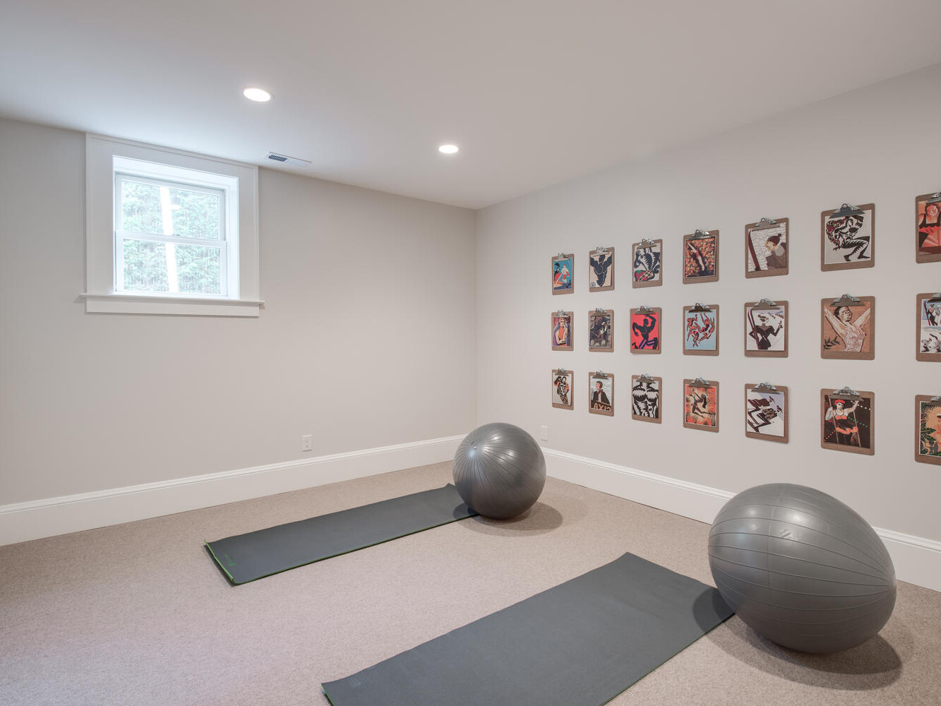 A minimalist room with two yoga mats on the floor, each with a large gray exercise ball. The walls are adorned with colorful art pieces. A small window lets in natural light, brightening the neutral-colored space.
