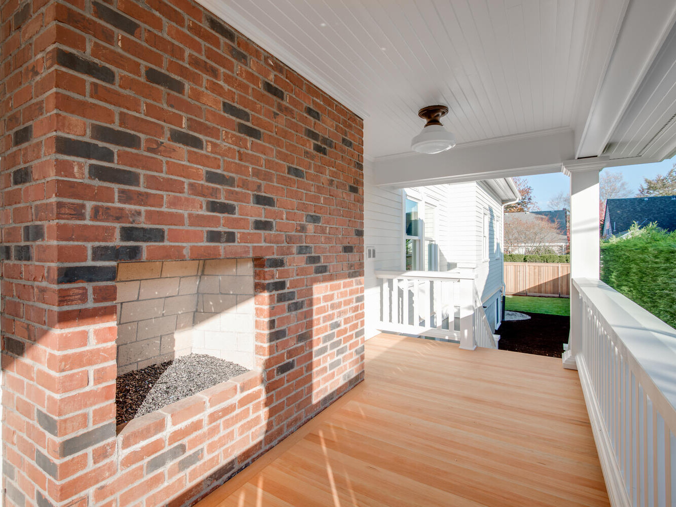 A bright porch featuring a brick fireplace without a chimney, white railing, and a polished wooden floor. The space has a white ceiling with a light fixture, and it overlooks a yard with green foliage and a wooden fence.