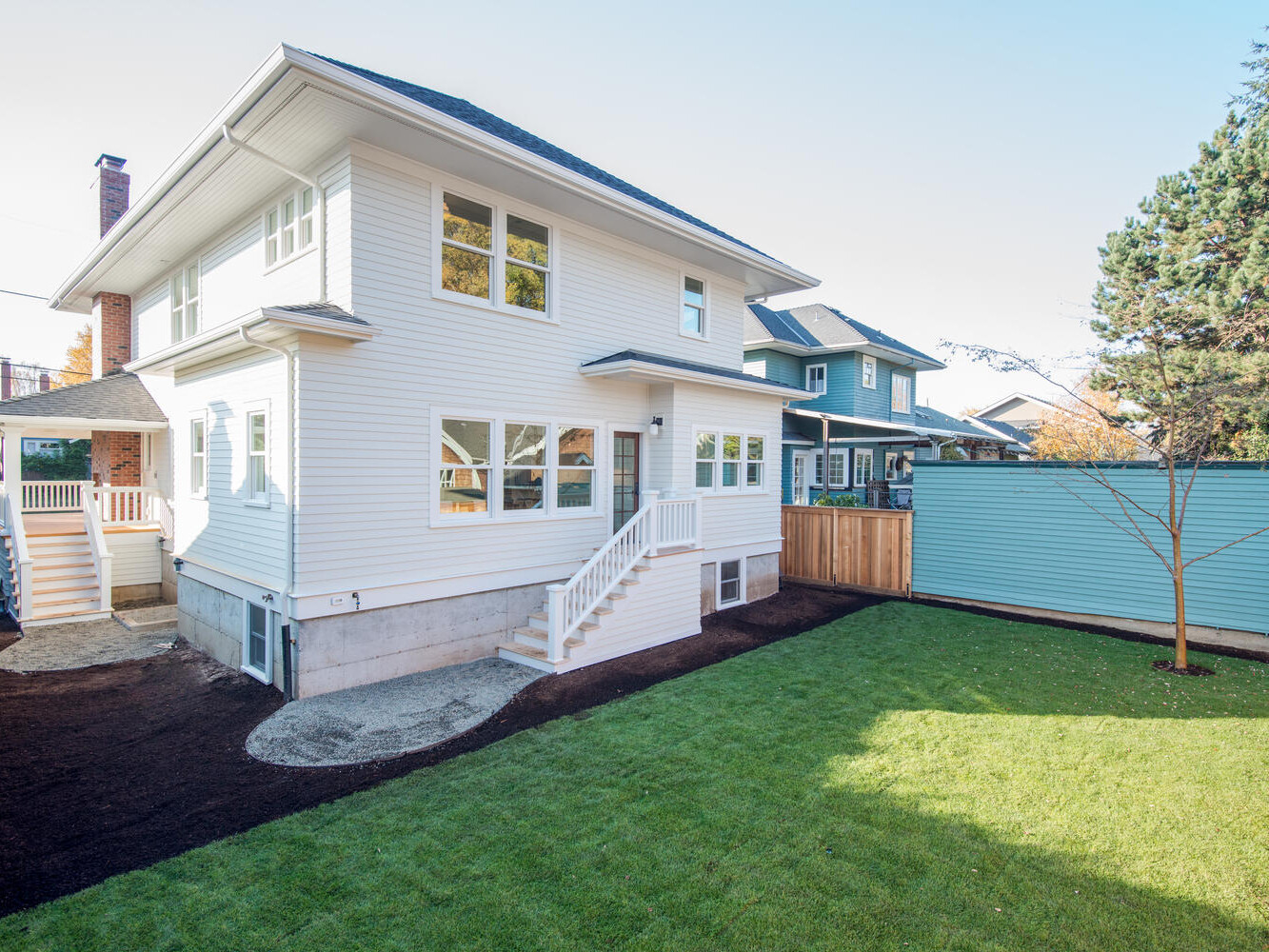 A two-story white house with large windows and a sloped roof is shown from the backyard. Theres a small concrete patio with stairs leading to a freshly landscaped lawn and a wooden fence, with neighboring houses visible in the background.