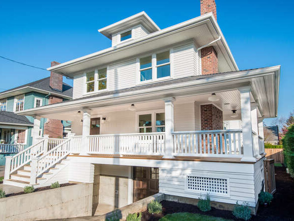 A two-story white house with a large front porch and chimney, featuring multiple windows. The house has a staircase leading to the entrance and a driveway with a garage door. The sky is clear and blue, adding to the bright atmosphere.