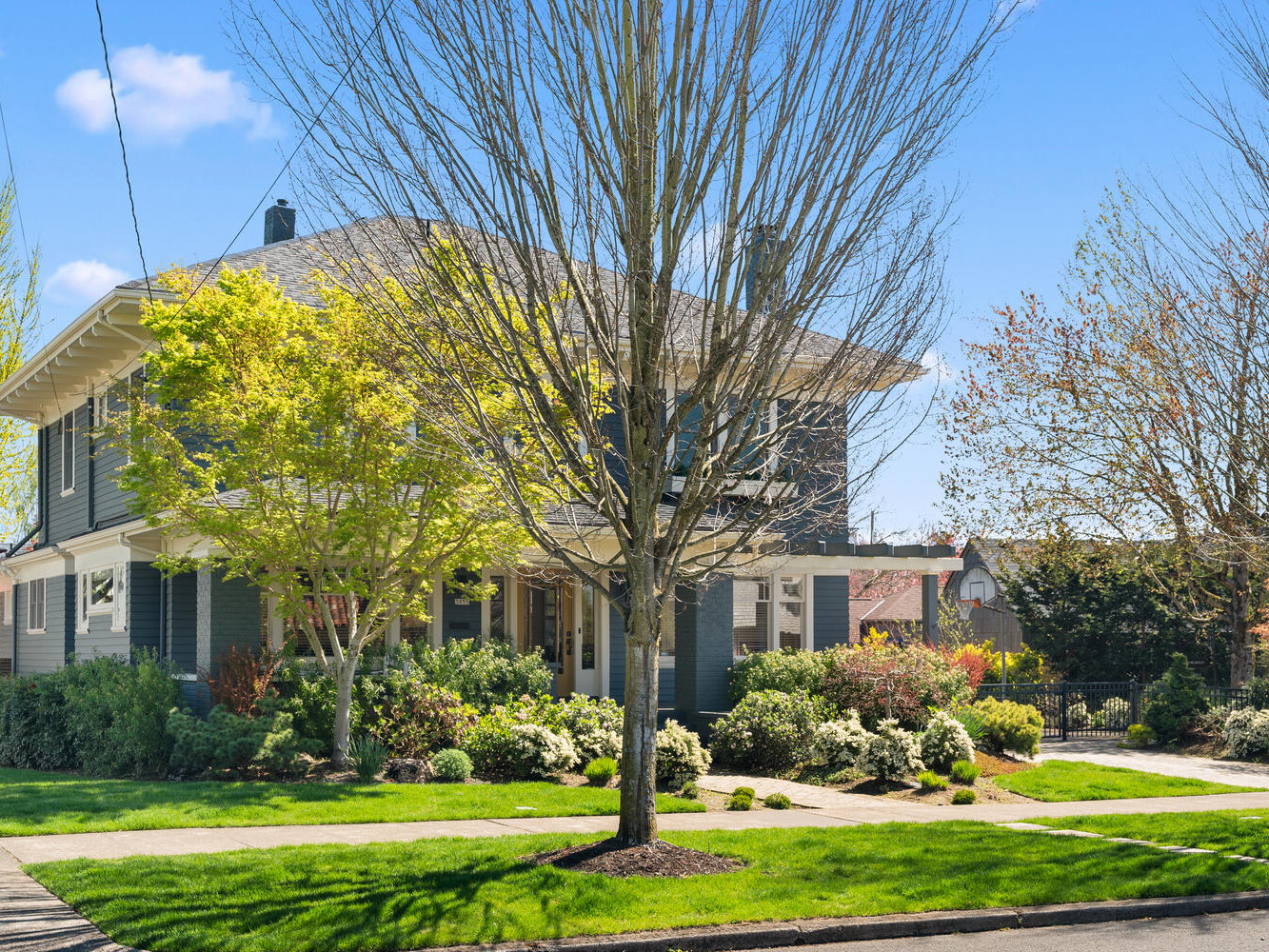 A suburban, two-story house with a gray exterior and a covered front porch, surrounded by well-maintained grass, trees, and shrubs. The sky is clear and blue, contributing to a bright and inviting atmosphere.