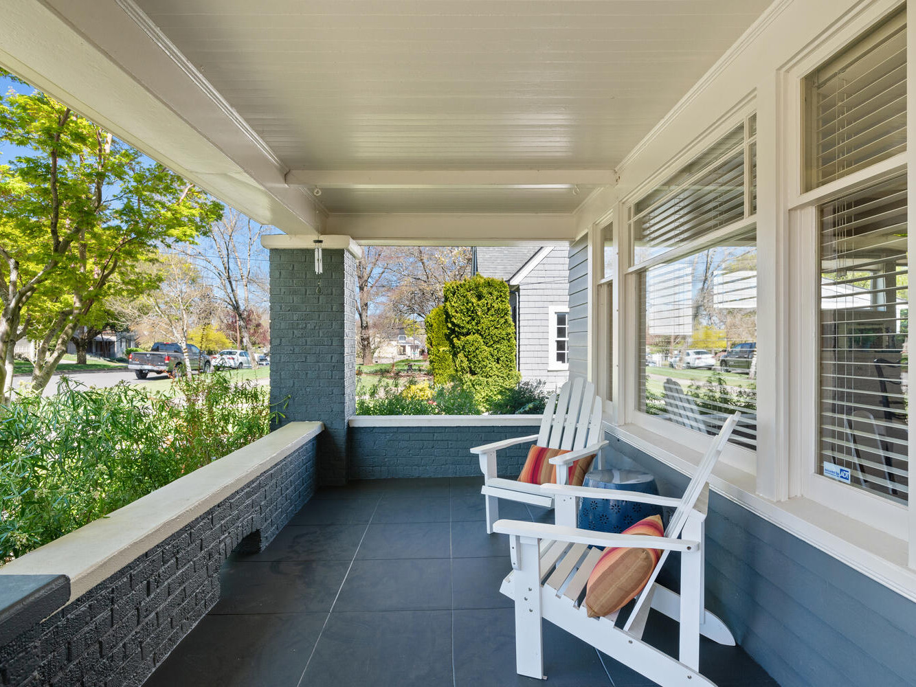 A cozy front porch with a white wooden bench and colorful cushions overlooks a residential street. The porch has light gray tiles and a partial brick wall. Lush greenery surrounds the area, and the house exterior is painted blue-gray.