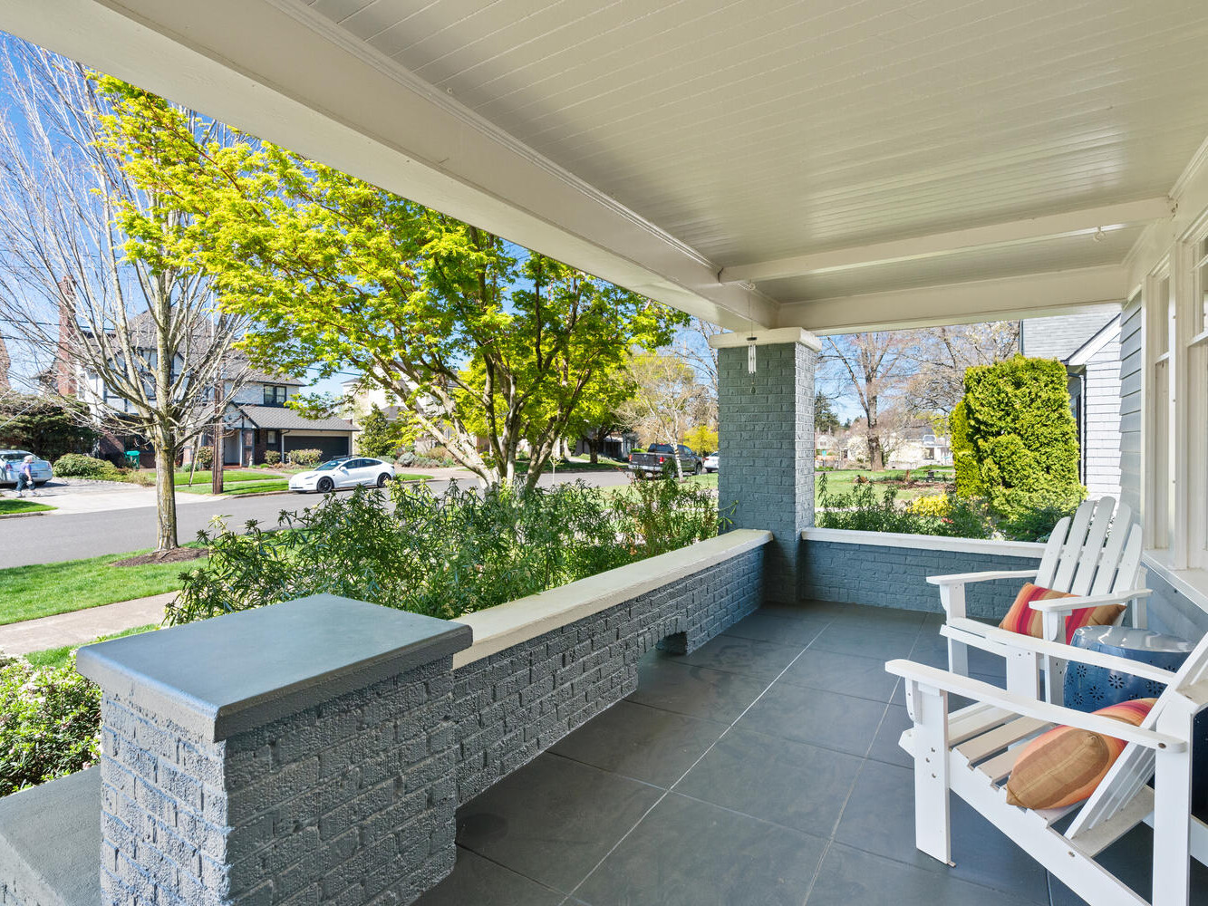 A cozy front porch with gray brick walls and white wooden ceiling. Two white Adirondack chairs with striped cushions face a leafy suburban street, showcasing a sunny day with clear skies and vibrant greenery.