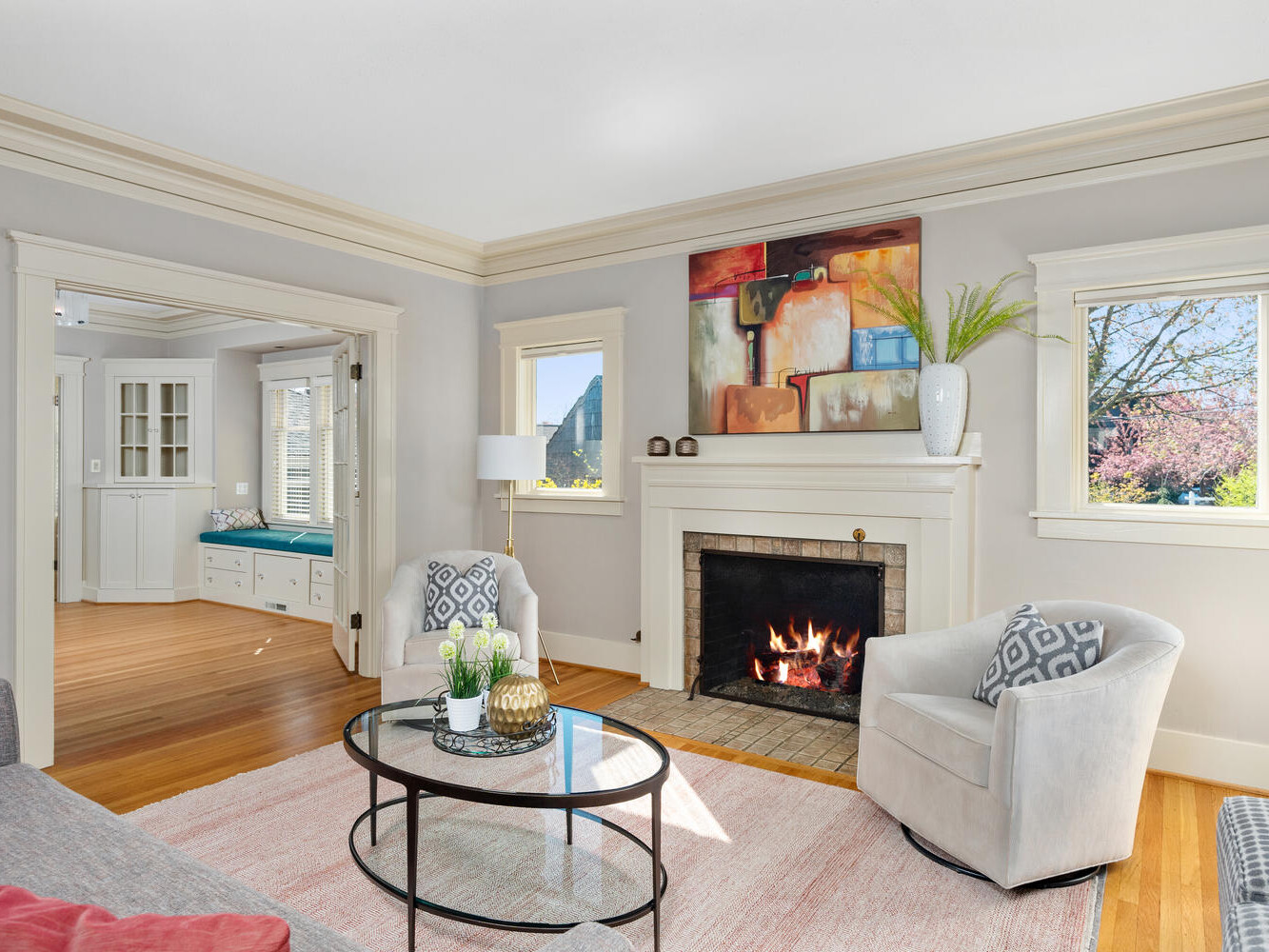A cozy living room features a lit fireplace, two armchairs, a round glass coffee table, and a gray sofa. An abstract painting hangs above the mantel. Large windows let in natural light, and a view into another room shows built-in cabinetry.
