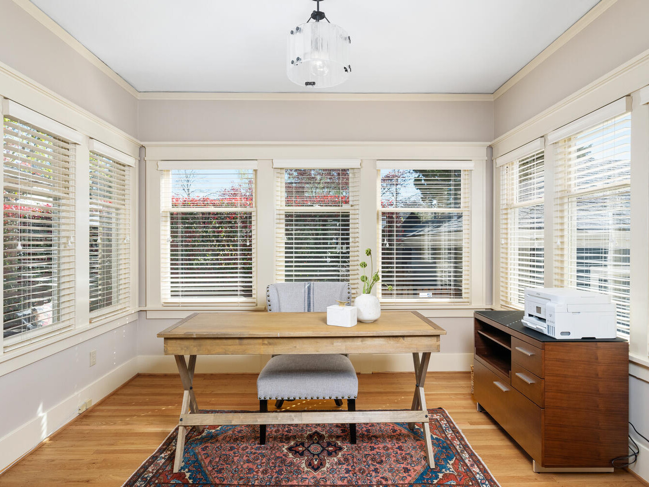 A bright home office with a wooden desk and cushioned chair. The room has large windows with white blinds, light walls, and a colorful rug on a wooden floor. A small wooden cabinet holds a printer and decorative items. A modern light fixture hangs above.