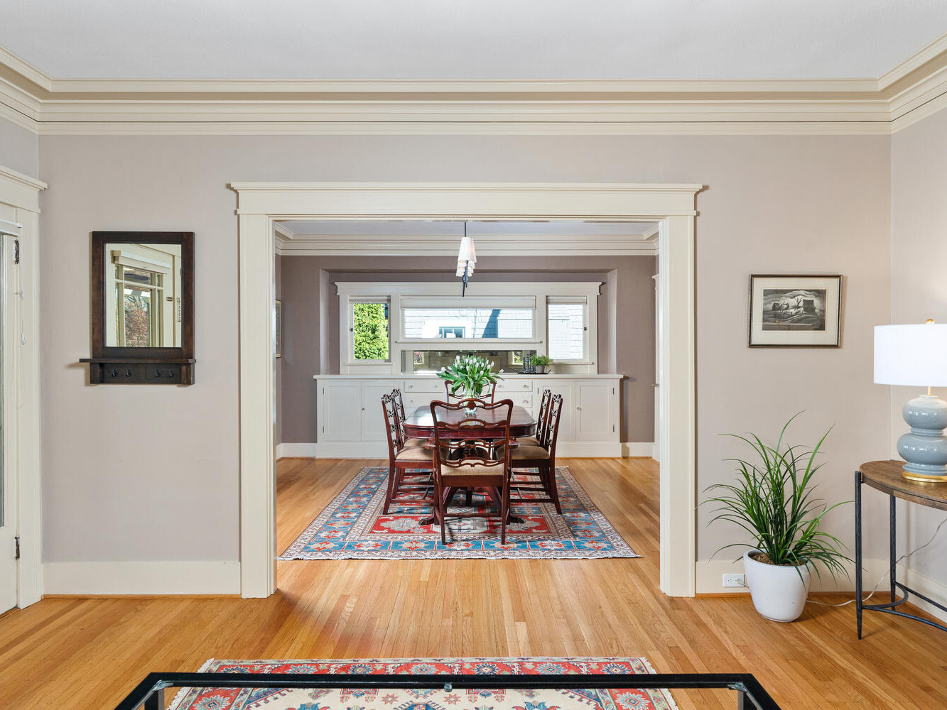 A dining room with a wooden table and eight chairs on a colorful rug. Natural light streams through windows. The room features hardwood floors, neutral walls, and decorative moldings. A plant and a lamp are on a side table.