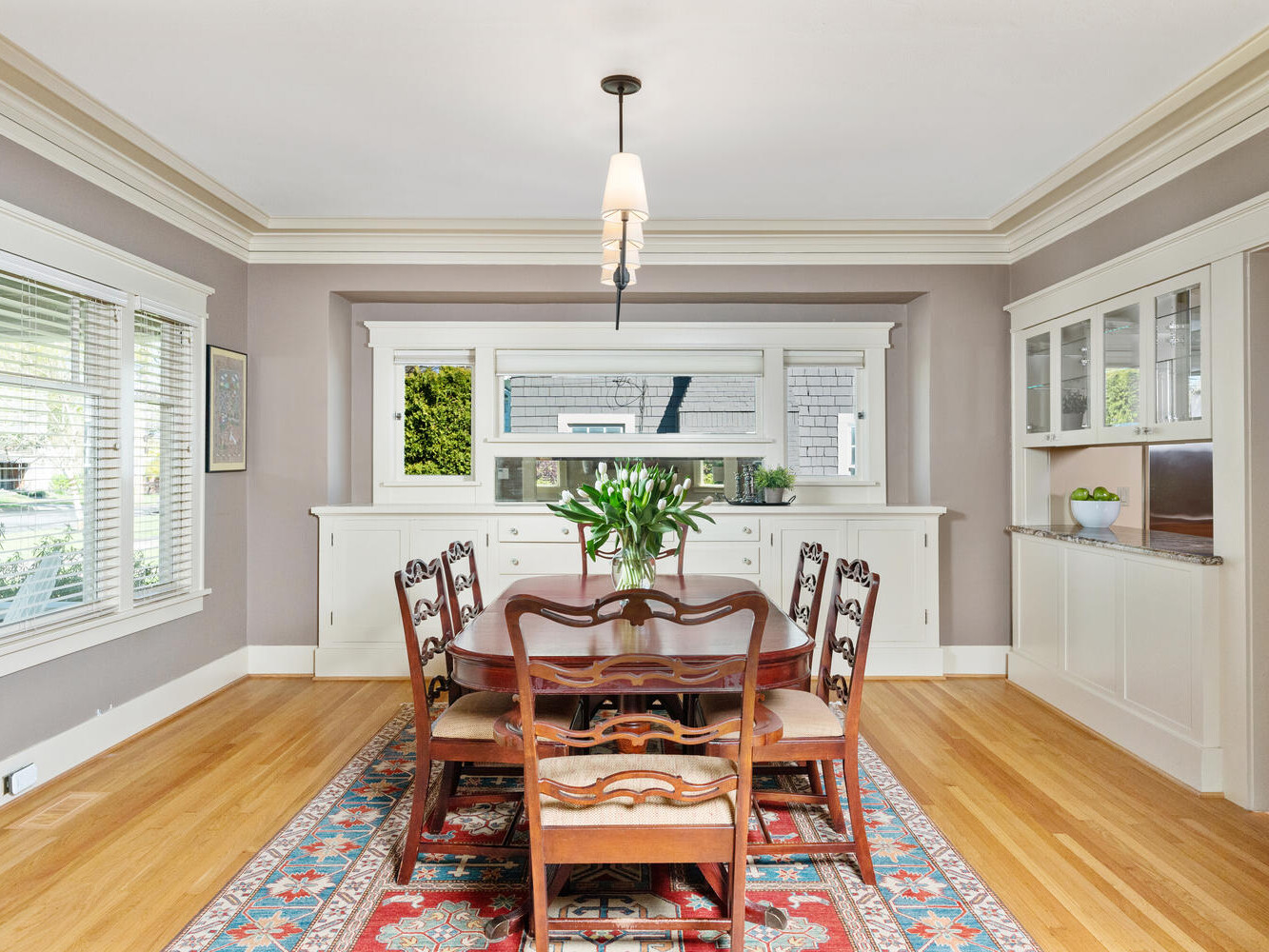 A traditional dining room with a wooden table and eight chairs on a patterned rug. A pendant light hangs above the table, and a vase with flowers is centered. White cabinetry lines the walls, with a window view to the outside.