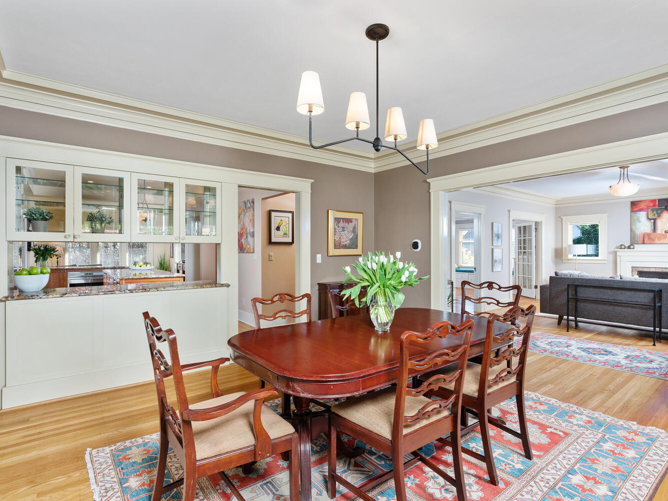 A well-lit dining room with a wooden table, six chairs, and a chandelier above. A cabinet with glass doors displays dishes. There are decorative rugs on the hardwood floor, and a vase with flowers on the table. A living room is visible in the background.