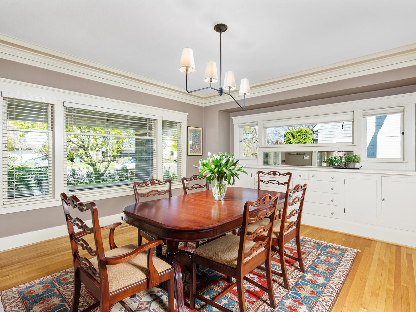 A classic dining room with a wooden table and six chairs on a patterned rug. A vase of tulips is centered on the table. The room has gray walls, white trim, large windows, and a built-in cabinet. A modern black chandelier hangs overhead.