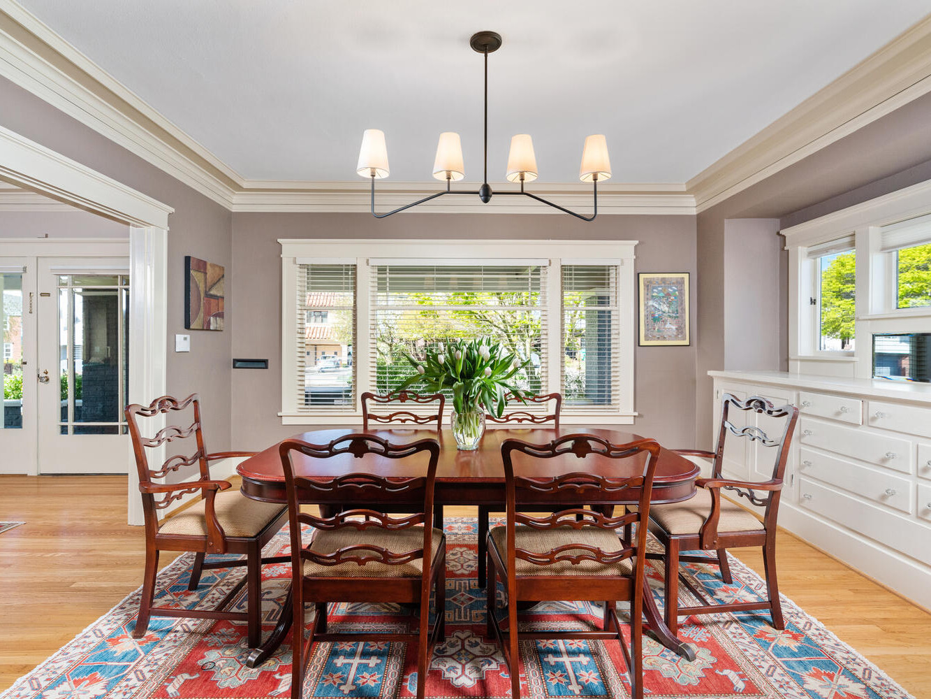 A dining room with a wooden table and six chairs, set on a colorful rug. A large chandelier hangs above the table, and a vase with flowers is in the center. The room has large windows allowing natural light.