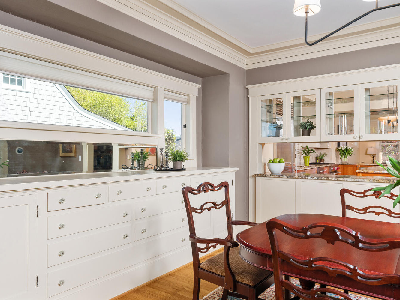 A bright dining room features a round wooden table with four chairs, a built-in sideboard, and a glass-front cabinet displaying dishes. Green plants decorate the surfaces, and a large window provides natural light and a view of the outdoor greenery.