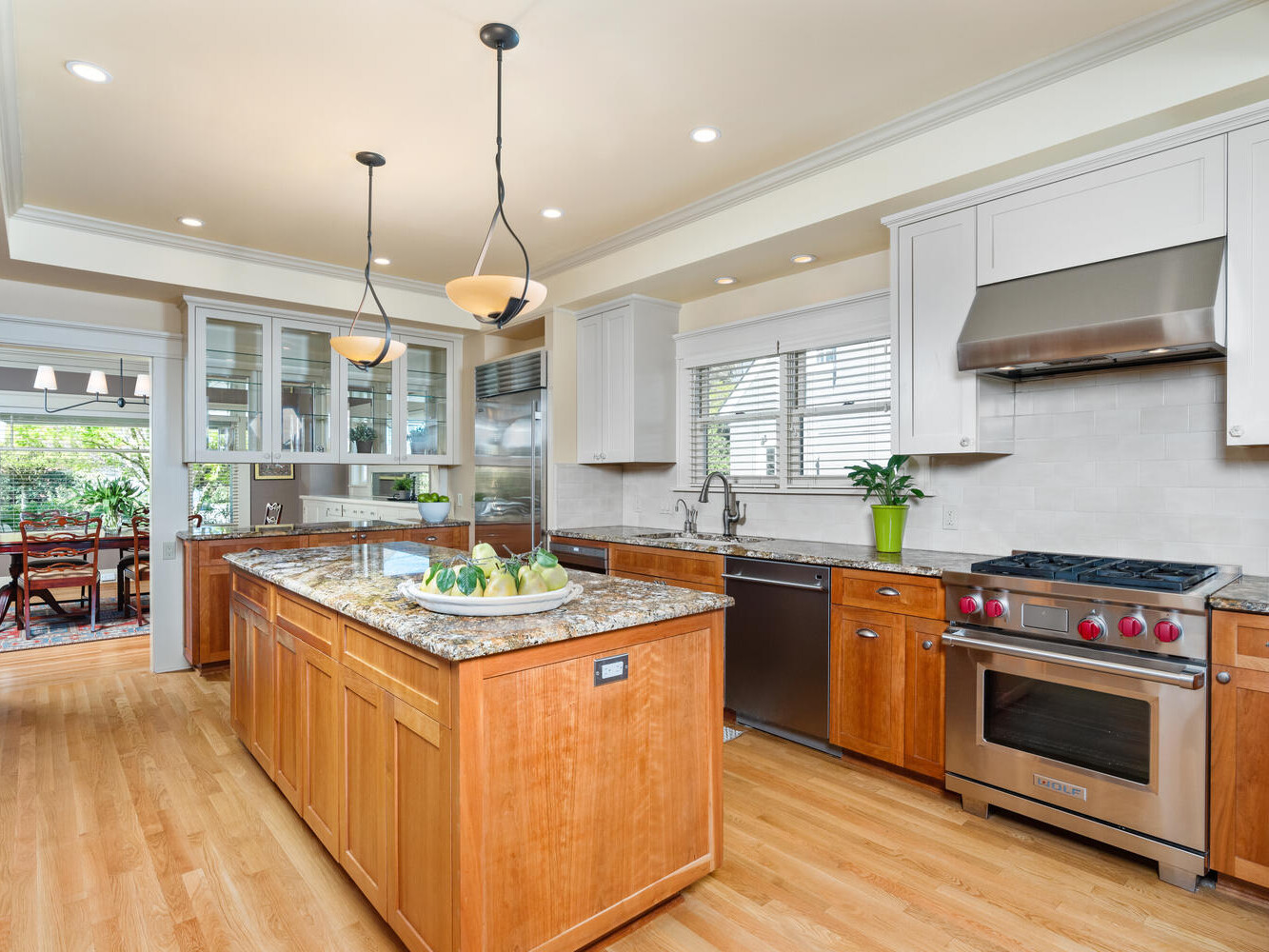 A spacious kitchen with wooden cabinets, stainless steel appliances, and a large island with a granite countertop. Pendant lights hang above the island, and a window offers natural light. A dining area is visible in the background.