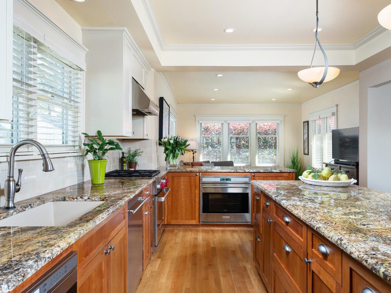 A spacious kitchen with wooden cabinets and granite countertops. A sink and dishwasher are on the left, while an oven is built into the cabinetry. A bowl of fruit sits on the island. Large windows let in natural light, illuminating the room.
