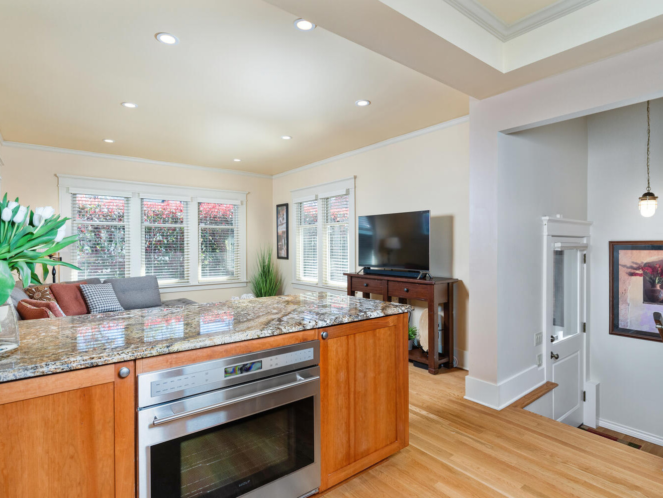 A bright, modern living room with wooden floors, a granite kitchen island, and an under-counter oven in the foreground. A sofa with decorative pillows, a TV on a wooden stand, and windows with blinds complete the cozy setting.