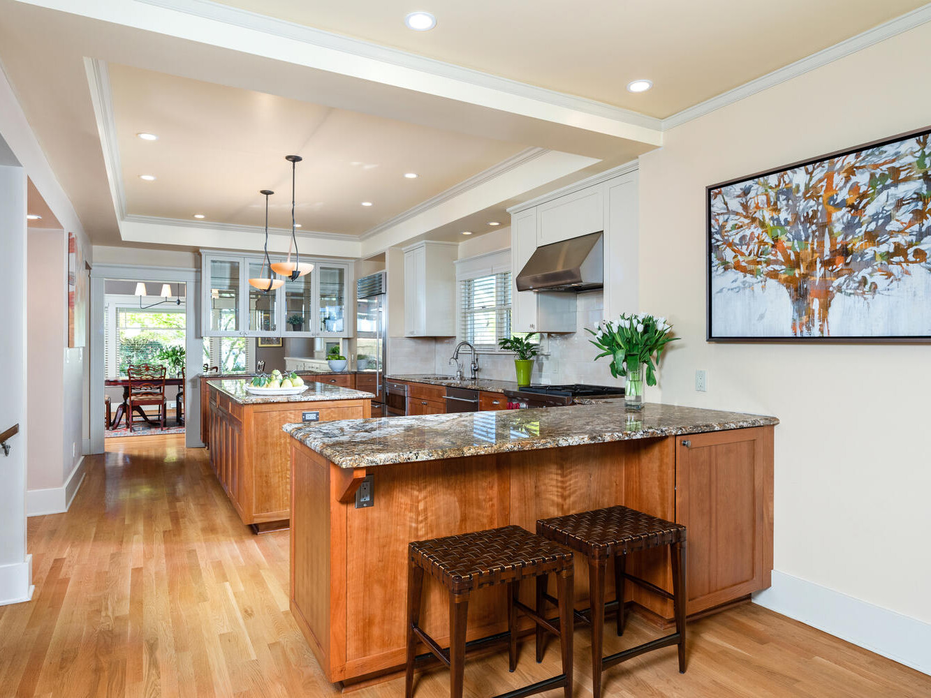 A modern kitchen with wooden cabinets, a granite island, and two wicker bar stools. Natural light streams in through large windows. A painting of a tree hangs on a cream-colored wall. Dining area visible in the background.