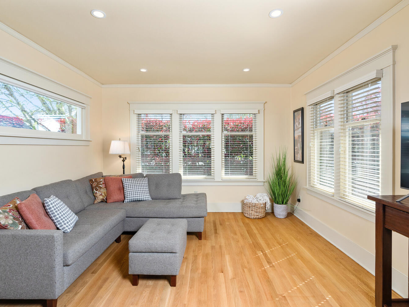 A bright, cozy living room with a large gray sectional sofa, colorful throw pillows, a small ottoman, and a flat-screen TV on a wooden stand. Large windows let in natural light, and potted plants add greenery. The wooden floor complements the warm decor.