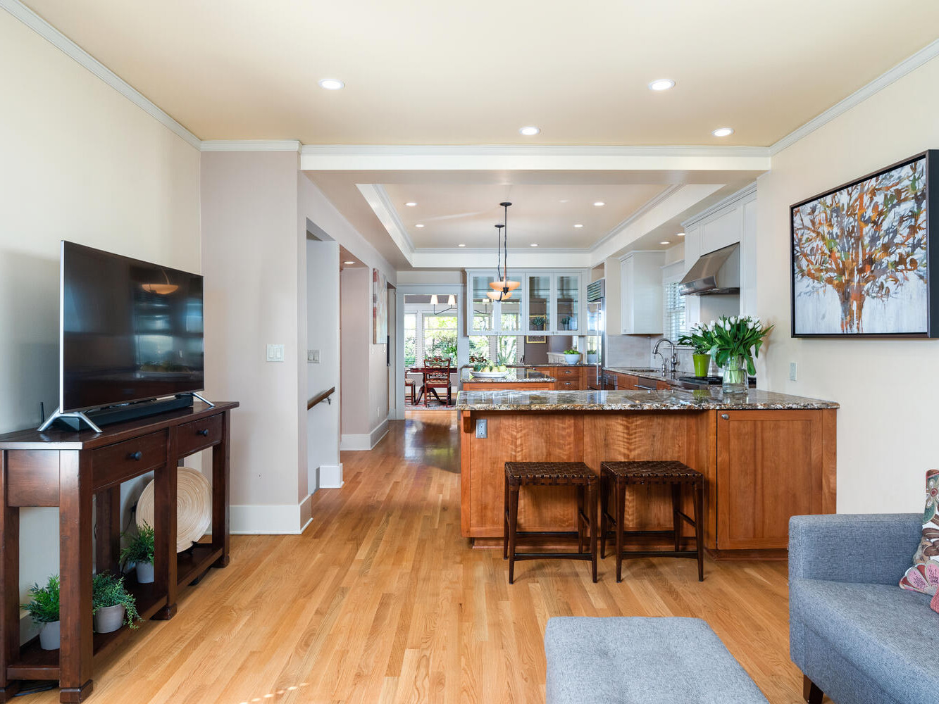 Modern living room with a flat-screen TV on a wooden console, hardwood floors, and an open kitchen with granite countertops. Cozy seating area in the foreground and a painting on the wall. Bright lighting and a view into the dining area.