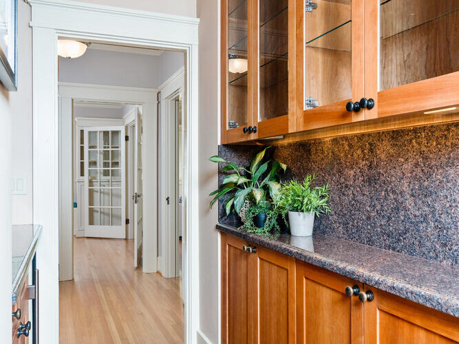 A hallway with wooden cabinets featuring glass doors on the right, topped with green plants. A red patterned rug covers the wooden floor. The hallway leads to white-framed double doors at the end.