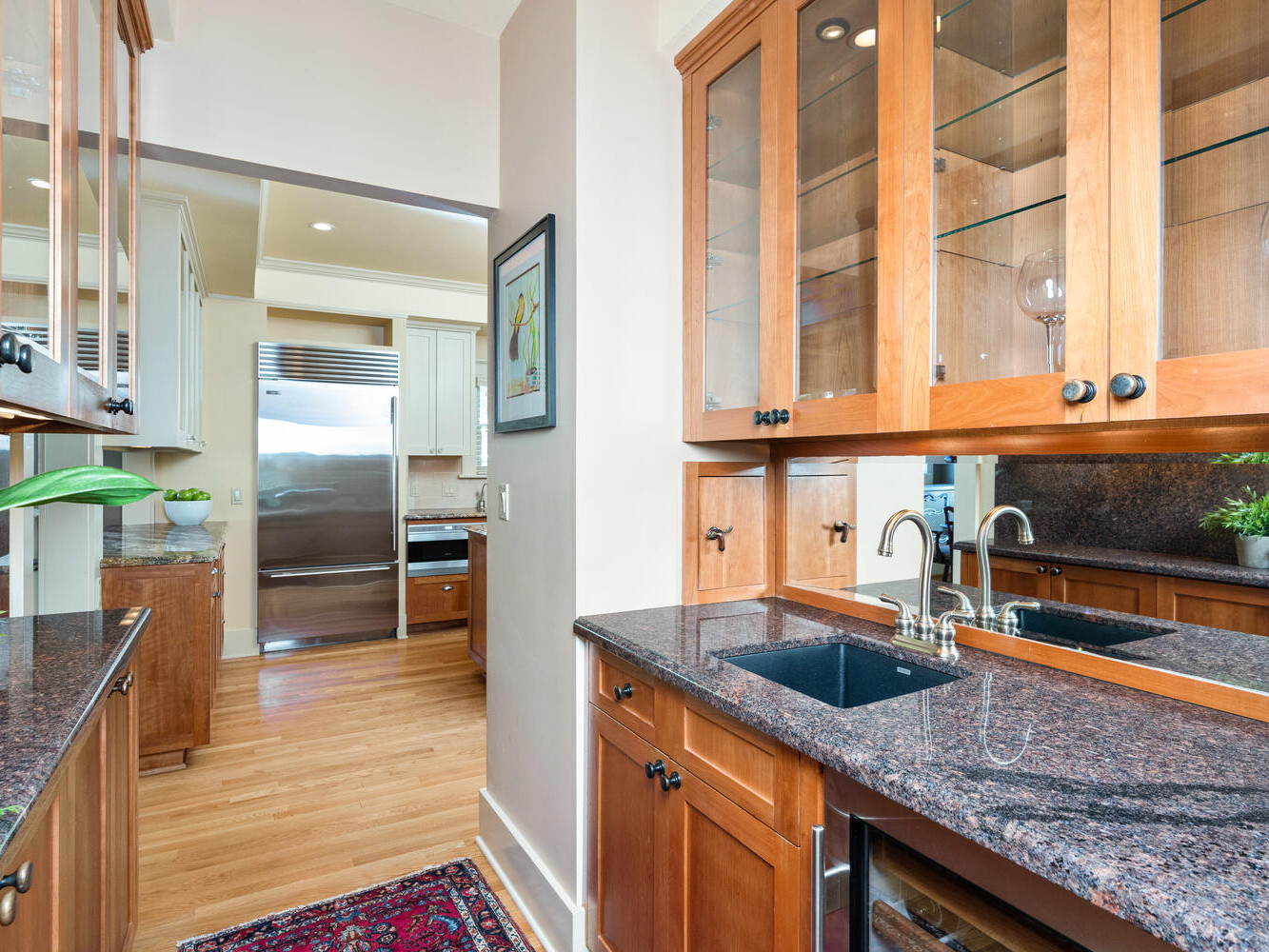 A kitchen with wooden cabinets, dark granite countertops, a sink, and glass-front upper cabinets. A red rug covers the wooden floor. The open area leads to a stainless steel refrigerator in the background. Green plants add a touch of nature.