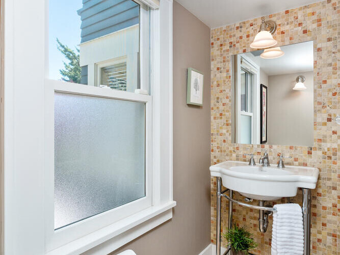 A small bathroom featuring a frosted window, a wall-mounted sink with a mirrored backsplash, a wall sconce above the mirror, and a tiled floor. A towel hangs near the sink, and a roll of toilet paper is mounted on the wall.