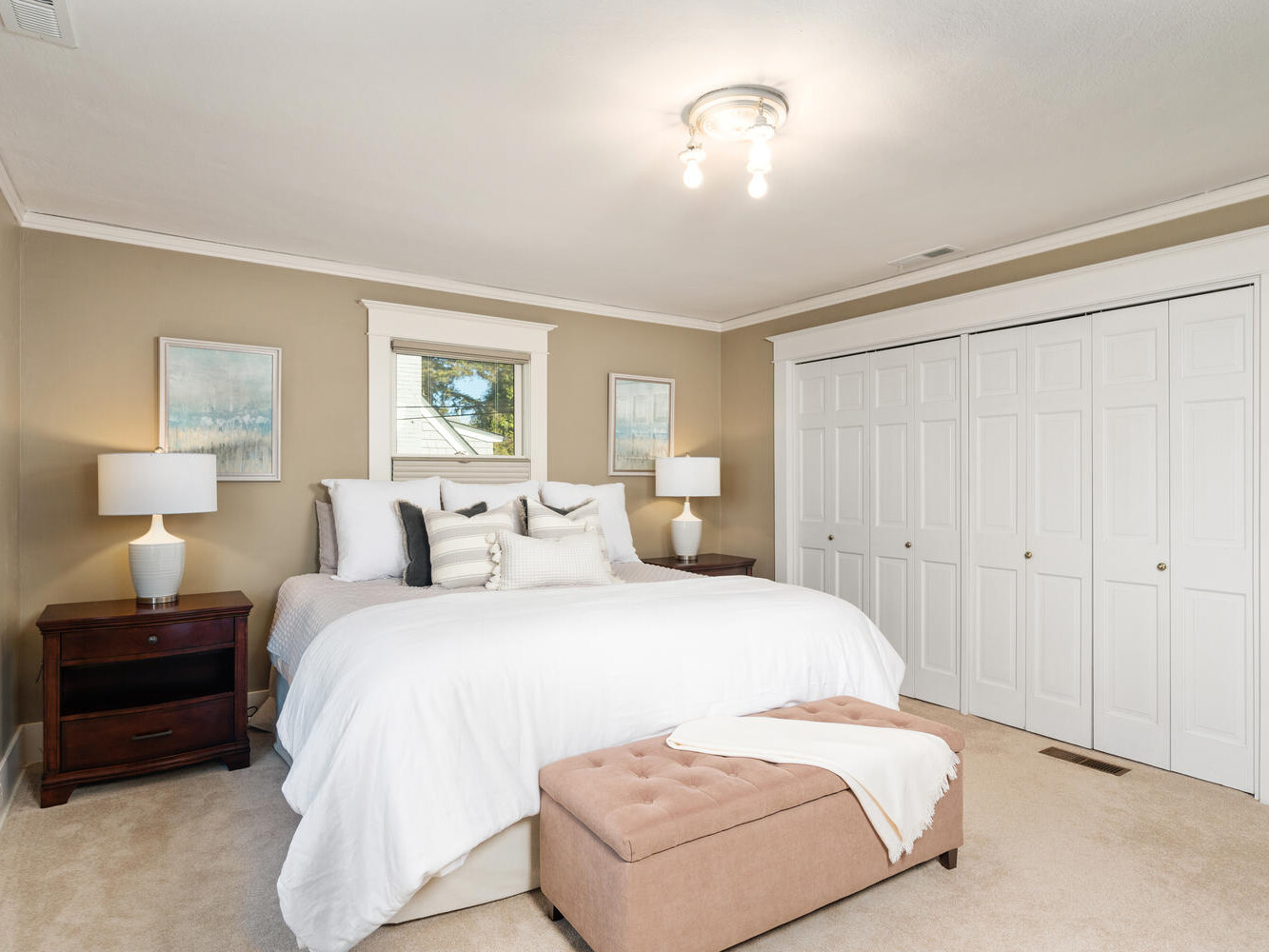 A cozy bedroom with a large bed featuring white bedding and multiple pillows. Nightstands with lamps flank the bed. Two framed pictures hang on the wall, and there is a cushioned bench at the foot of the bed. A closed closet is visible.