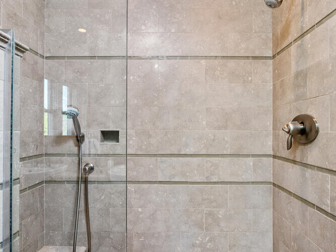 A modern shower with gray stone tiles, featuring a glass partition. Equipped with a fixed showerhead and a handheld shower on a sliding bar. A recessed niche is built into the wall, and a small step can be seen on the left side.