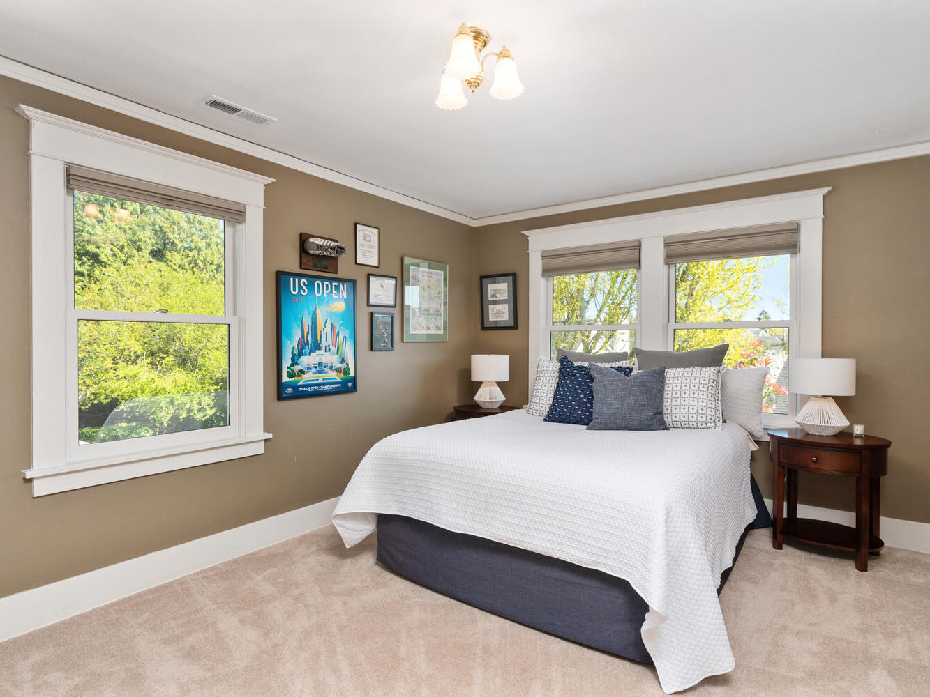 A bedroom with beige walls and cream carpet features a neatly made bed with white and blue pillows. Art and a bright poster adorn the walls. Two windows offer views of trees and greenery. A small bedside table with a lamp is on the right.